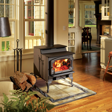 Black wood stove with fire burning in it . firewood nexted to it and windows behind it. other room that is scene has tables and chairs