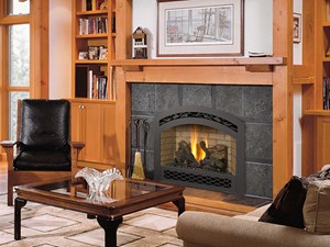 black fireplace insert with fire burning with brown hearth and bookselves. Chair and couch with table in a room.