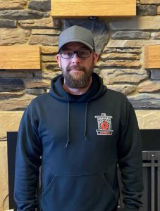 man in a black sweatshirt and grey baseball hat. Has glasses and a gotee